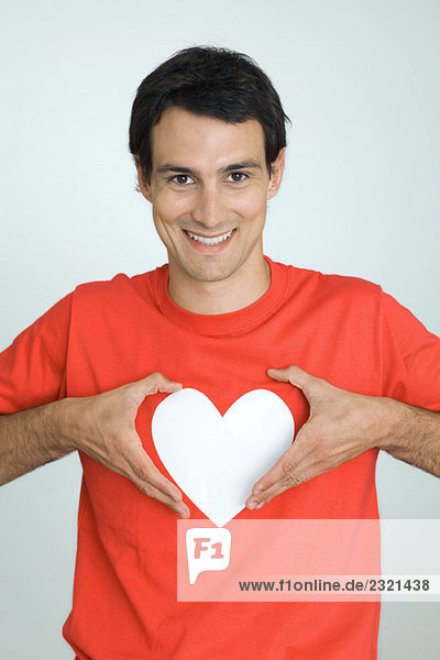 Mann im T-Shirt mit Herzsymbol  hält Hände um das Herz  lächelt die Kamera an.