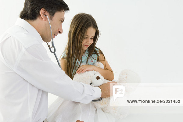 Arzt sitzt mit kleinem Mädchen  untersucht Teddybär mit Stethoskop