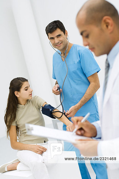 Krankenpfleger  der den Blutdruck eines jungen Patienten misst  lächelt in die Kamera  Arzt schreibt auf die Zwischenablage im Vordergrund.