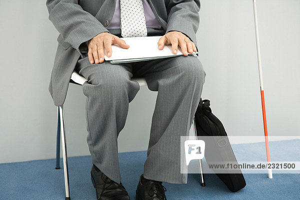 Visually impaired man sitting in chair  holding documents on lap  cropped view  chest down