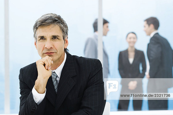 Graying businessman with hand under chin  looking away  colleagues chatting in background