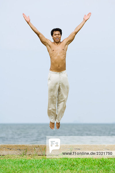 Mann springt mit erhobenen Armen in die Luft  lächelt in die Kamera  das Meer im Hintergrund