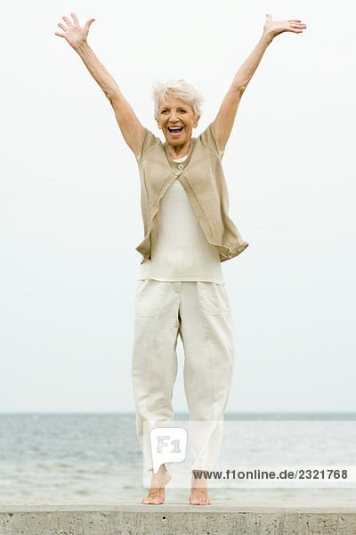 Seniorin auf Zehenspitzen am Strand stehend  Arme erhoben  lächelnd