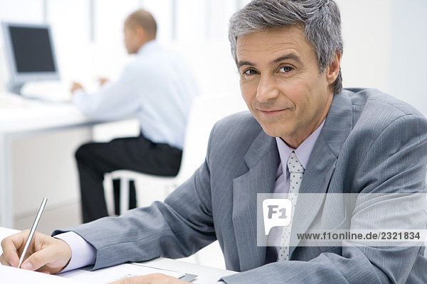 Businessman sitting at desk  using pen  smiling at camera