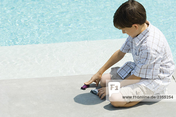 Boy sitting on ground  playing with toy cars  next to pool