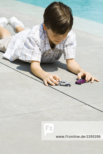 Boy lying on ground  playing with toy cars  next to pool