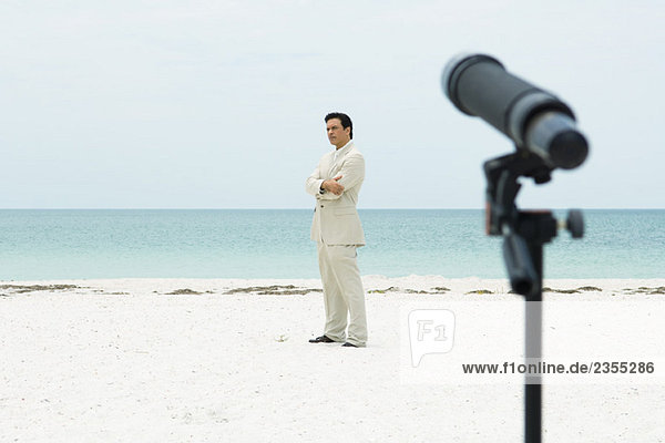 Geschäftsmann am Strand stehend mit gefalteten Armen  Teleskop auf ihn gerichtet im Vordergrund