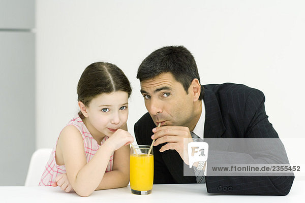 Kleines Mädchen und Vater trinken aus einem Glas Saft mit Strohhalmen  Blick in die Kamera