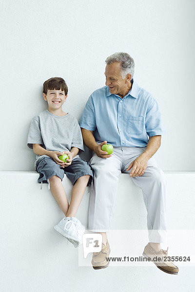 Grandfather and grandson sitting side by side on ledge  holding apples  both smiling
