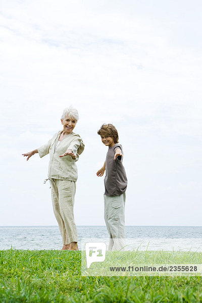 Großmutter und Enkel stehen im Freien mit ausgestreckten Armen  beide lächelnd vor der Kamera  Ozean im Hintergrund