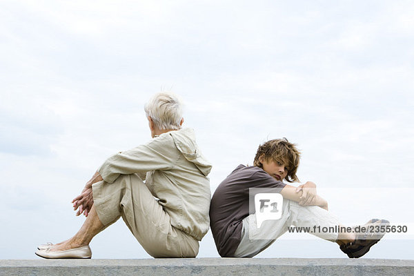 Großmutter und Enkel sitzen Rücken an Rücken  Frau schaut weg  Junge schaut in die Kamera