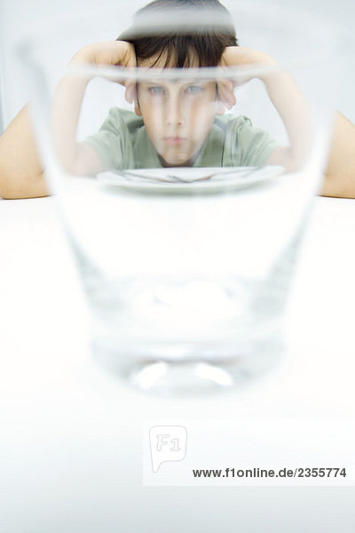 Junge schmollt am Tisch  schaut durch das Wasserglas in die Kamera.