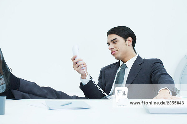Young businessman sitting at desk with feet up  looking at landline phone  smiling