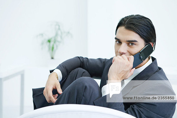 Young businessman sitting with knees up  using cell phone  looking at camera