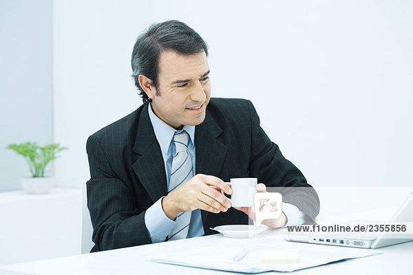Erwachsener Geschäftsmann am Schreibtisch mit einer Tasse Kaffee  lächelnd
