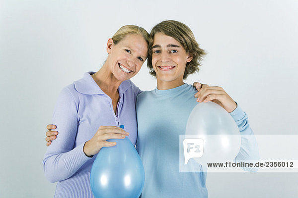 Mutter und jugendlicher Sohn mit den Armen um die Schultern  mit Luftballons  lächelnd vor der Kamera.