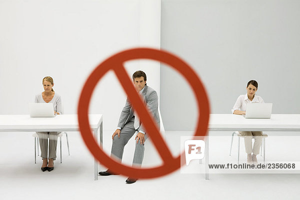 Two women seated in front of laptop computers  warning sign over man between them  man looking at camera
