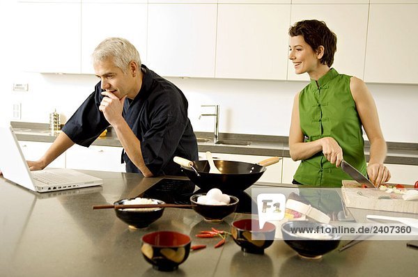 Ein reifer Mann mit einem Laptop und einer erwachsenen Frau  die in der Küche Gemüse hackt.
