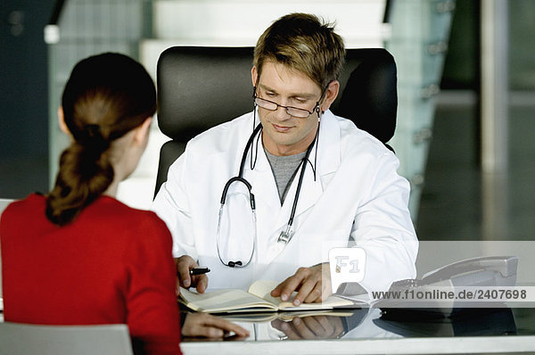 Ein männlicher Arzt sitzt mit einer Patientin und macht sich Notizen in seinem Büro.