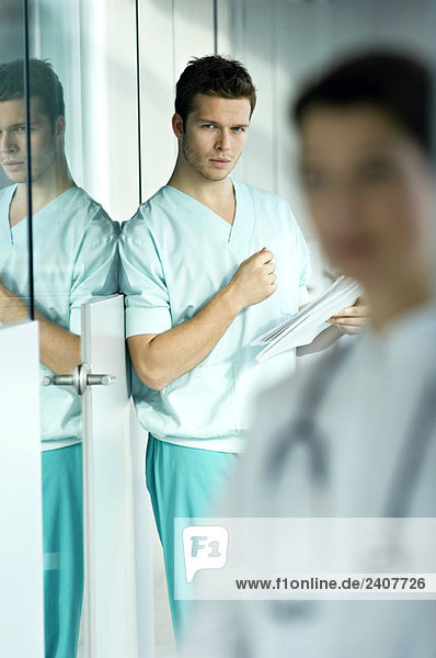 Portrait eines männlichen Arztes  der eine Krankenakte hält und sich an eine Wand lehnt.