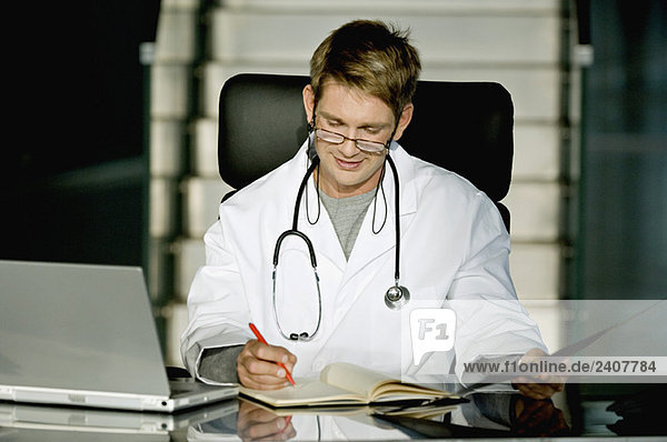 Männlicher Arzt sitzt an einem Schreibtisch in seinem Büro und schreibt in ein Notizbuch.