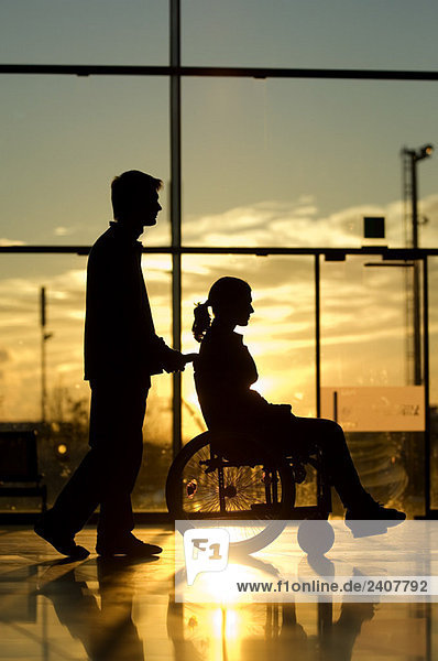 Silhouette eines Mannes  der eine Patientin im Rollstuhl schiebt.