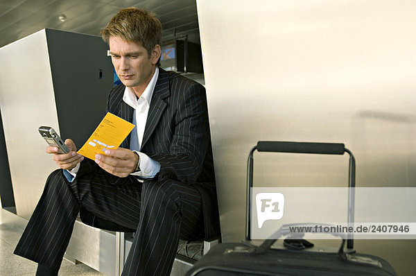 Geschäftsmann  der auf einem Flughafen sitzt und ein Mobiltelefon bedient.