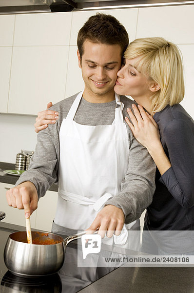 Junger Mann beim Kochen und eine junge Frau  die ihn von hinten in der Küche küsst.