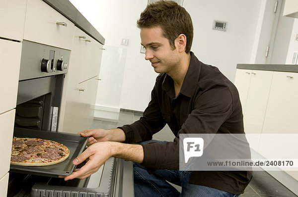 Junger Mann stellt ein Tablett mit Pizza in den Ofen und lächelt.