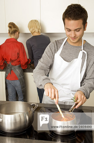 Junger Mann beim Kochen und zwei junge Frauen  die hinter ihm in der Küche stehen.