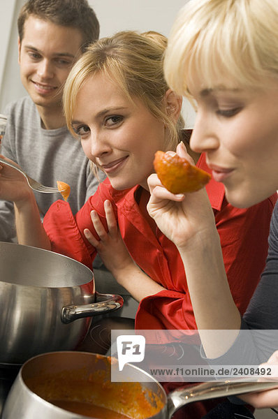 Junge Frau probiert Tomatensauce mit einer Schöpfkelle mit einem jungen Mann und einer jungen Frau neben ihr in der Küche.