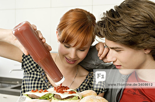 Young woman pouring tomato sauce on burgers with a teenage boy standing beside her