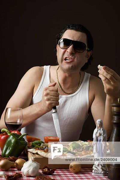 Stereotypischer Italiener  der ein Messer in ein Schneidebrett steckt und Pizza isst.