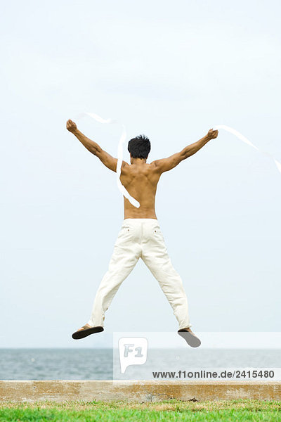 Mann springt in die Luft mit Luftschlangen in den Händen  Rückansicht  Ozeanhorizont im Hintergrund