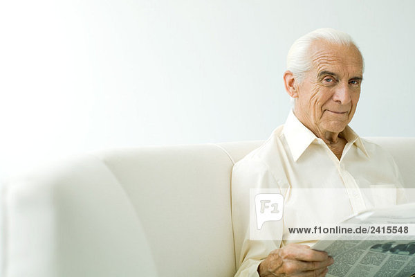 Senior man sitting on sofa  holding newspaper  smiling at camera