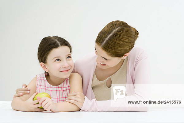 Frau am Tisch sitzend mit Arm um die Schulter der Tochter  lächelnd  Apfel haltendes Mädchen