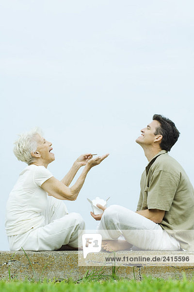 Seniorin und erwachsener Sohn sitzen von Angesicht zu Angesicht auf dem Boden  teilen sich Essen zum Mitnehmen  lachend