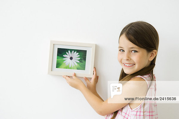 Kleines Mädchen hält gerahmtes Bild an der Wand  lächelt über die Schulter vor der Kamera