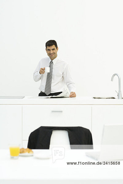Mann steht an der Küchentheke mit Kaffee in der Hand und lächelt in die Kamera.