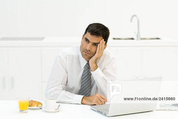 Businessman sitting in kitchen with breakfast and laptop  leaning head on hand  looking away