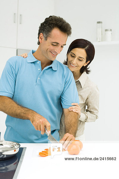 Couple standing together in kitchen  man slicing tomato  both smiling