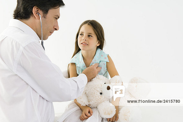 Kinderarzt hört Mädchenherz mit Stethoskop  Mädchen hält Teddybär