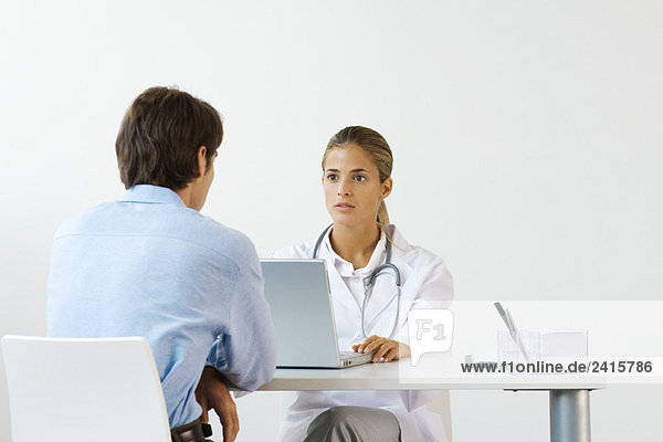 Female doctor sitting across from male patient at desk  using laptop computer