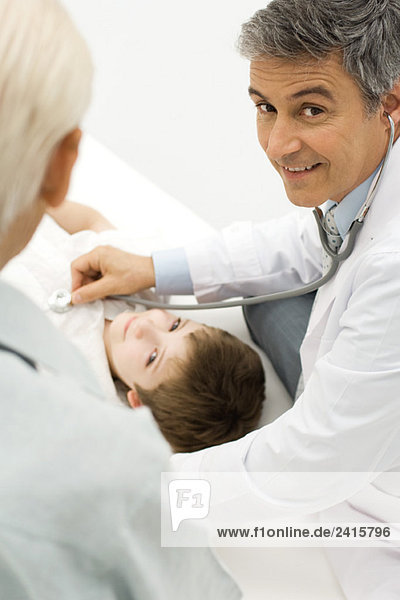 Arzt hört auf das Herz des jungen Patienten  lächelt über die Schulter vor der Kamera