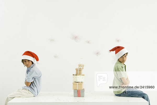 Zwei Brüder sitzen Rücken an Rücken mit gefalteten Armen  Weihnachtsgeschenke gestapelt zwischen ihnen  ein Blick auf die Kamera