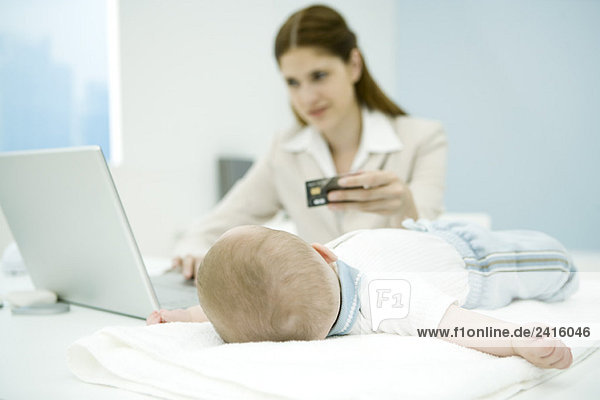 Junge berufstätige Mutter beim Kreditkartenkauf  Baby auf dem Schreibtisch liegend  Fokus auf Baby im Vordergrund