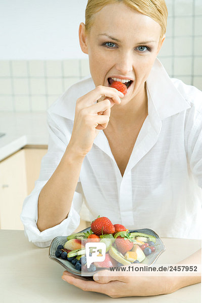 Woman holding bowl of fruit salad  eating strawberry  looking at camera