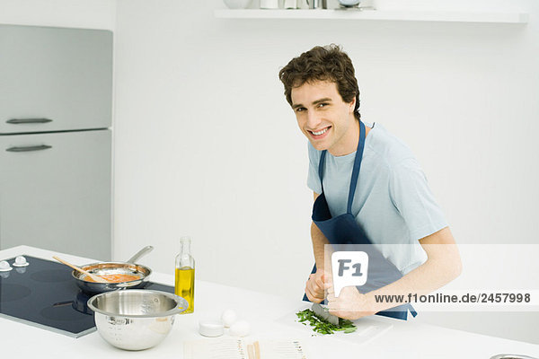 Junger Mann zerkleinert Basilikum in der Küche  lächelt in die Kamera.