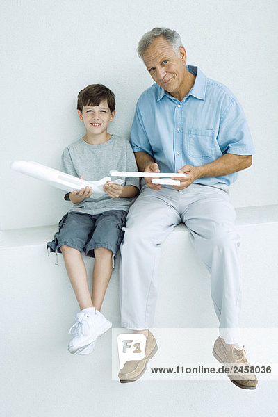 Grandfather and grandson sitting side by side  holding model airplane pieces  smiling at camera