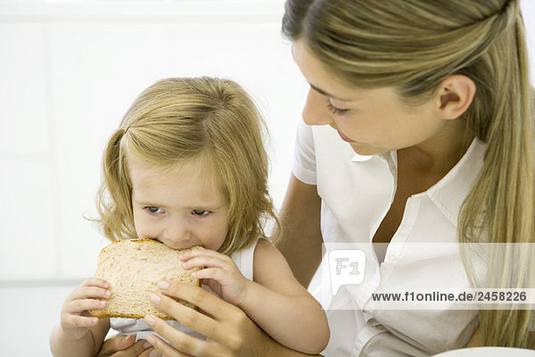 Kleines Mädchen sitzt auf dem Schoß ihrer Mutter und isst eine Scheibe Brot.